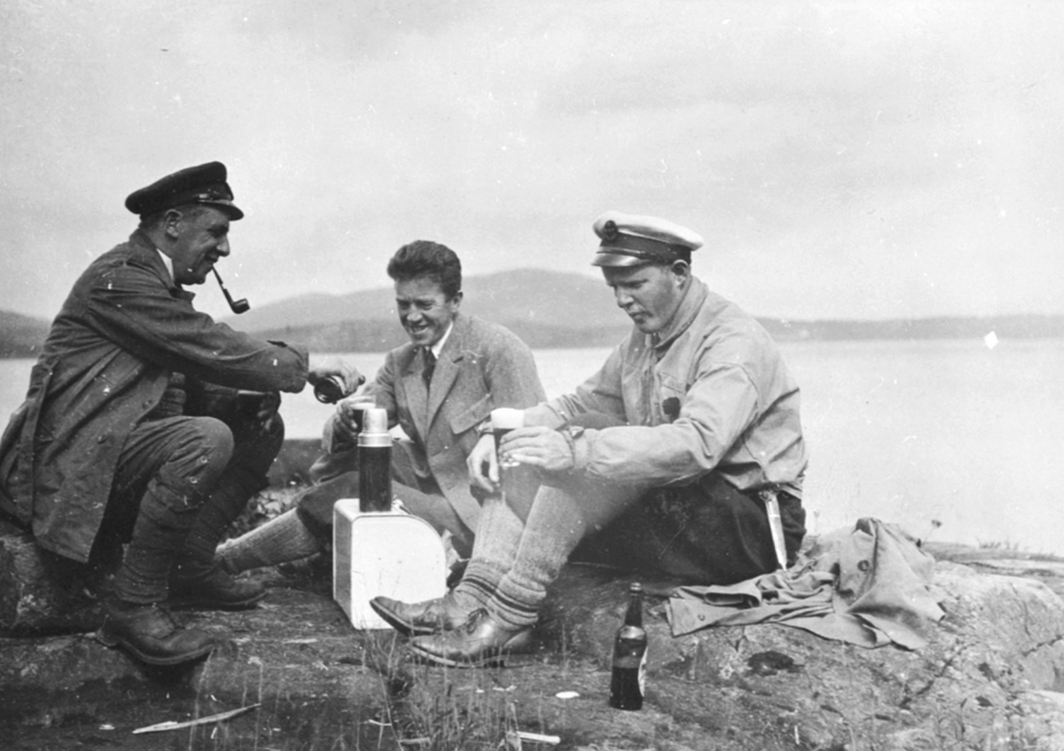 Tre menn raster i strandkanten. Fra venstre er Nickelsen, Bjørnerud, tannlege Karl Løken med skyggelue og slirekniv. Flasker og termos.