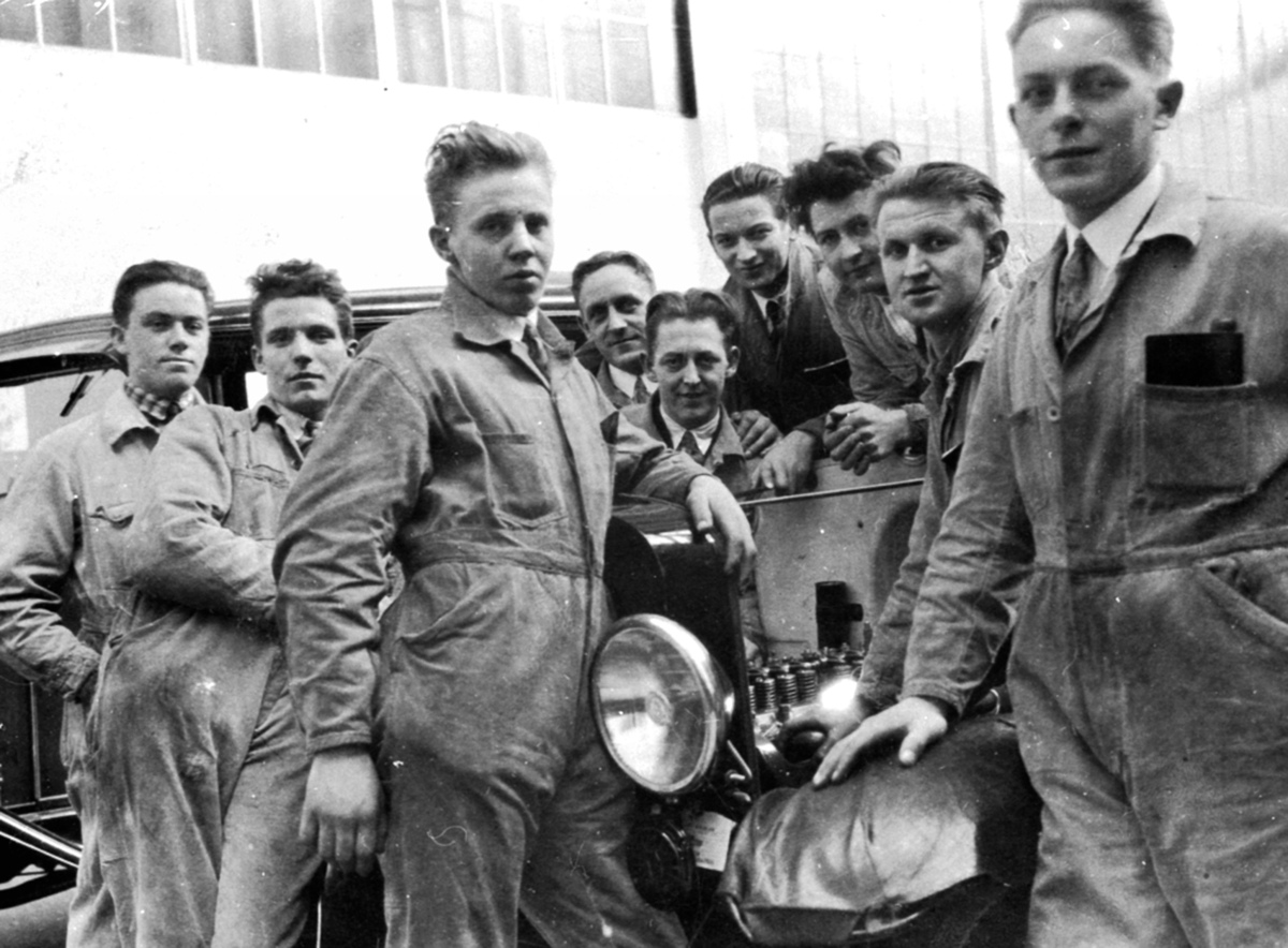 Gruppe menn i kjeledress rundt bil. Bilmekanikere på kurs. Johannes Haug (1909-1986) fra Nes BIlverksted.