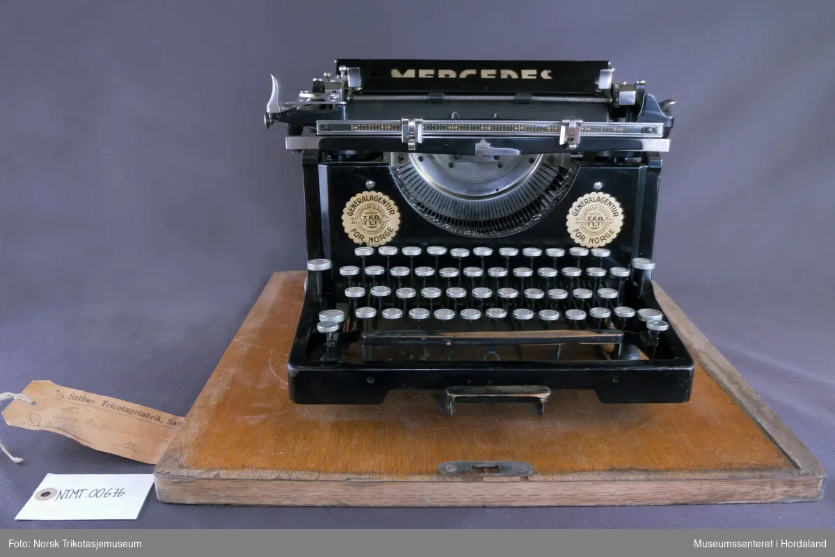 Mercedes standard skrivemaskin med gammelt tastatur, men utan bokstaven "å". Pica skrift, 9-tommers valse (kan kun skrive A4). Skrudd fast på treunderlag og har medfølgjande metallokk: ei transportabel standardmaskin.