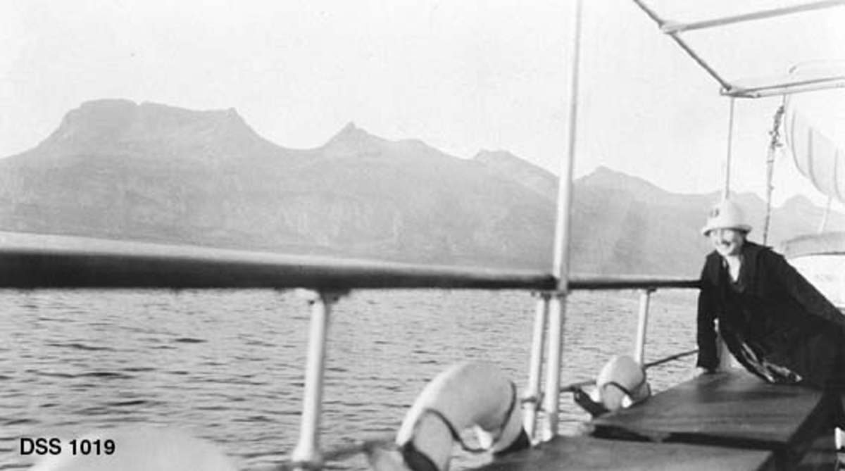 Fjellkjeden De sju søstre sett fra sørøst.  Fotografiet er tatt fra et båtdekk, der det sitter ei kvinne med mørk kåpe og lys hatt på en benk ved rekka og ser ut over sjøen. 