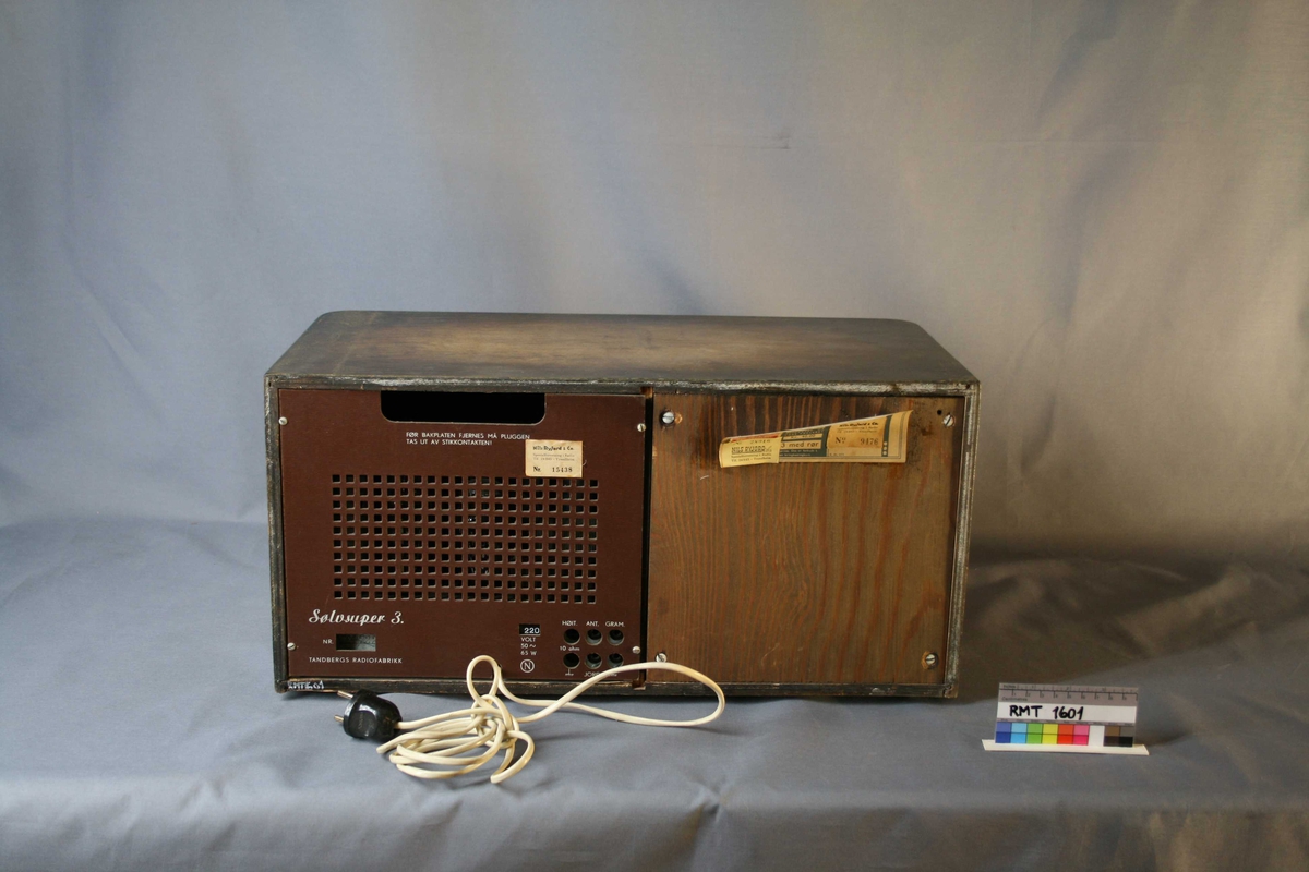 Rektangulær brun radio. Forside: høyttaler med tekstiltrekk, skala og fire funksjons-, og søkeknapper. Bakside: bakplate og strømuttak.