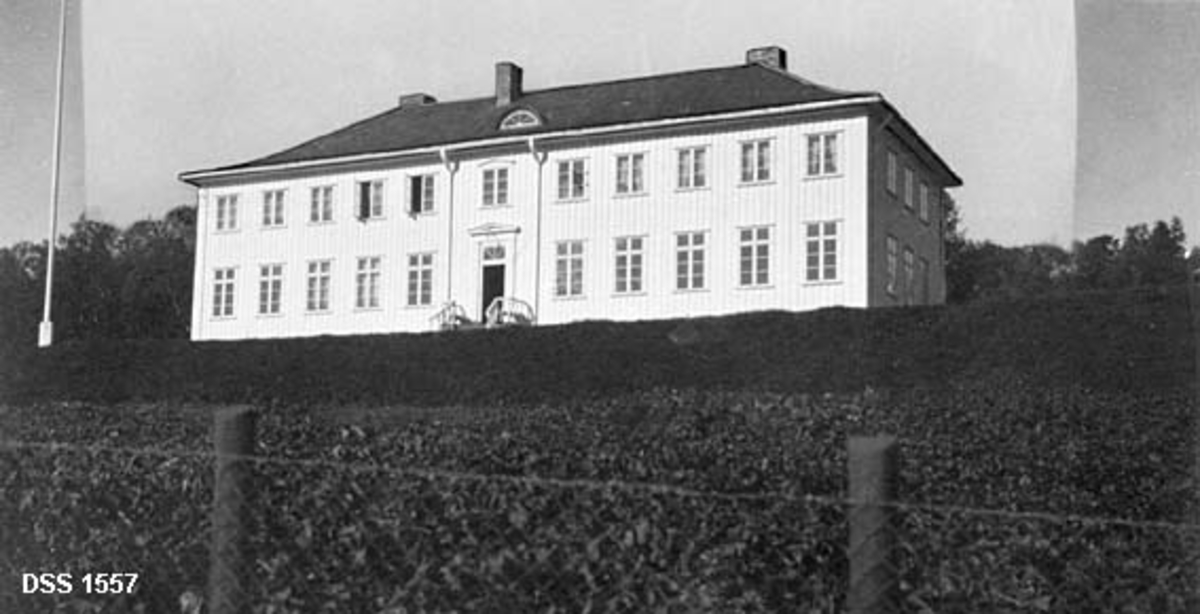 Undervisningsbygningen ved Steinkjer skogskole i Nord-Trøndelag, fotografert i 1926, cirka to år etter at den sto ferdig.  Det dreier seg om en stor, toetasjes bygning, utført i tretoms lafteplank, som ble isolert med papp og bordkledd på begge sider.  De utvendige fasadene ble tidlig kvitmalte.  Valmtaket ble tekket med skifter.  Bygningen står på en bakkekam og har et klassisk strilpreg med dør og trapp midt på fasaden mot hagen nedenfor.  I forgrunnen et nettinggjerde på stolper.  Taket er noe retusjert, og himmelen er "forhøyet" på dette fotografiet. 


Litt mer informasjon om Steinkjer skogskole og bygginga av den nye skogskolebygningen i perioden 1922-1924 finnes under fanen «Opplysninger».