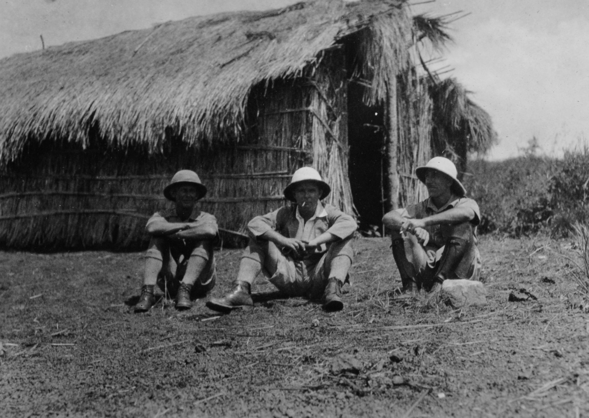 Tre menn sitter på bakken utenfor en strå-/bambushytte i Uganda.