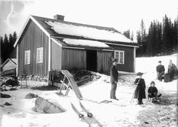 Ukjent familie foran et hus, ei jente på ski og en gutt på k