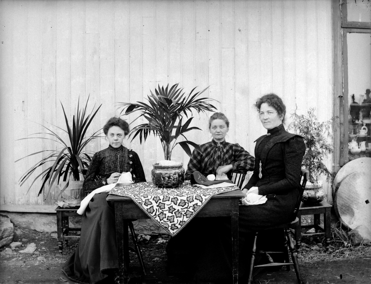 Tre kvinner driver med håndarbeid, syr, ved en husvegg. 