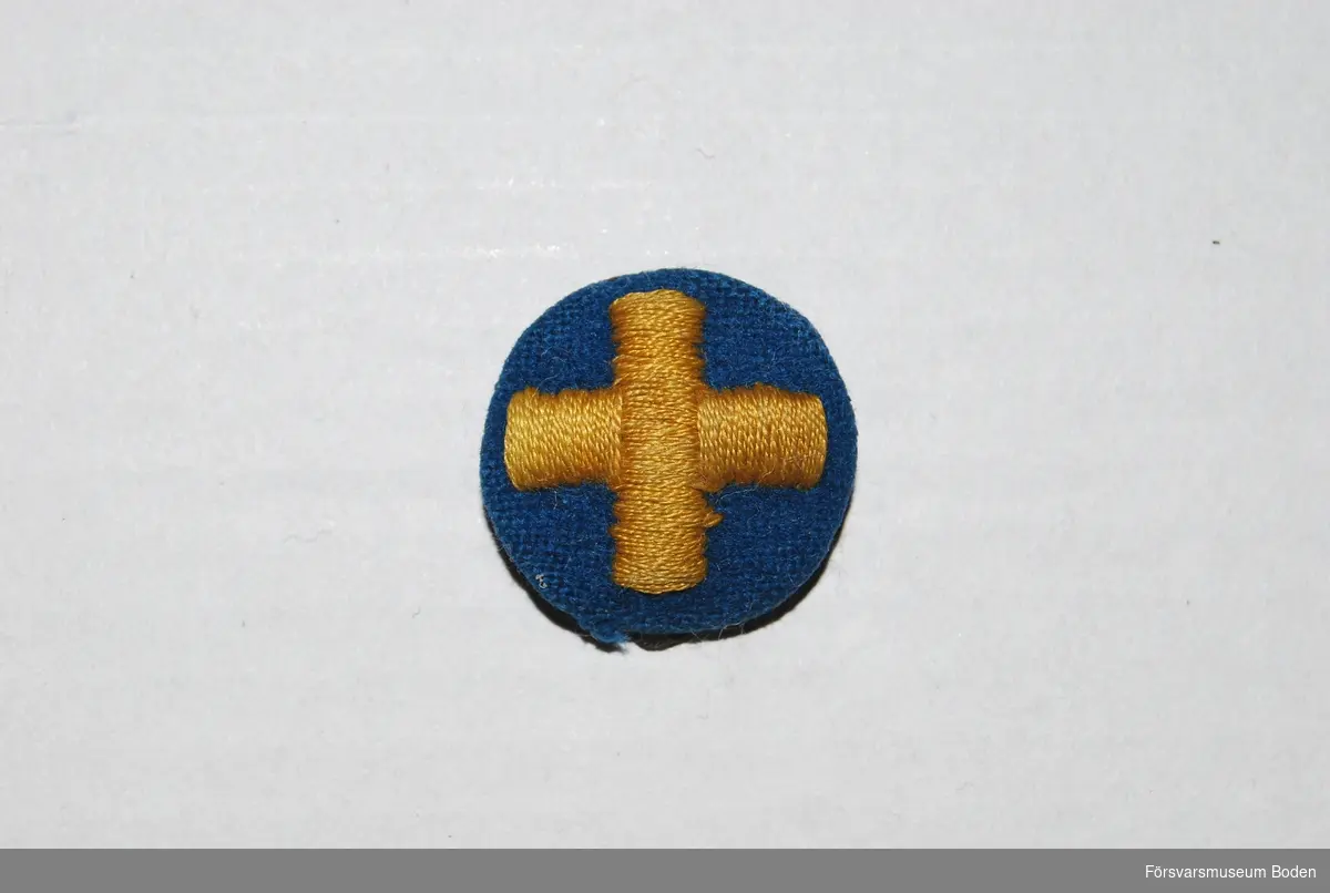 Kors broderat med gul silkestråd på rund matta av mellanblått kläde. För modellexemplar, se mössmärket registrerat under AM.070494.
