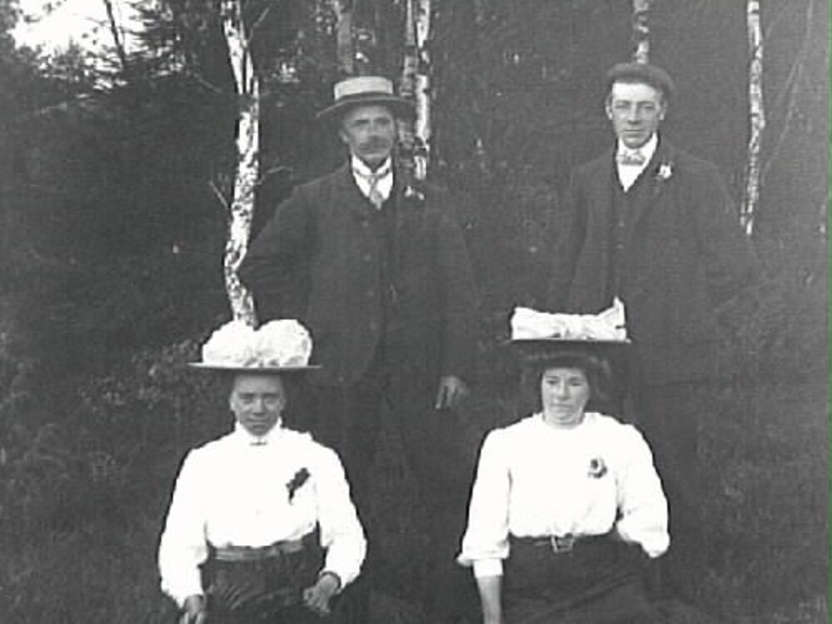 Gruppbild av två kvinnor och två män fotograferade i en skogsbacke. Kvinnorna bär stora hattar. Stående från vänster Edvard Johansson, Klas Johansson. Sittande från vänster Emma och Hulda Johansson.