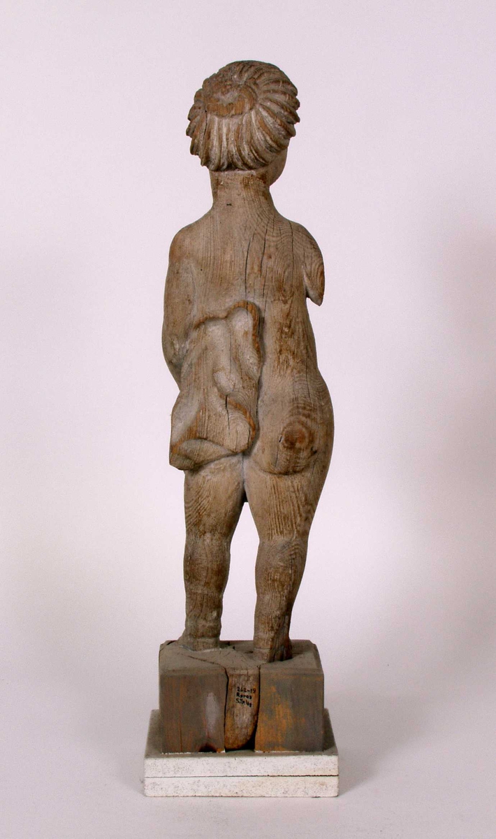 Utskåret trefigur. Det finnes rester av hvit maling i fordypninger (anatkelig linoljemaling. Dertil finnes det et tynt gråhvitt malingslag over hele overflaten. Dette lag er sekundært ettersom det også er påført der deler mangler.