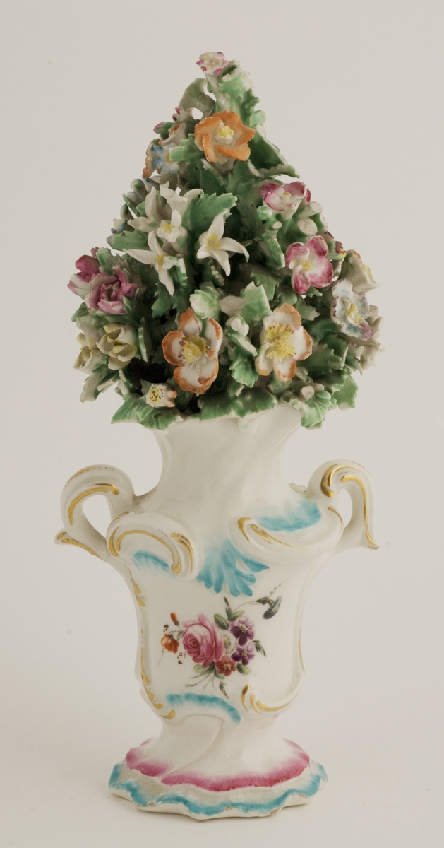 Urne med lokk. Porselen, lokk rikt dekorert med blomster som går opp i en topp. To hanker med plastisk naturalistisk blomsterbukett. Usignert. 
Lokket går opp i en topp. Rikt dekorert medblomster.