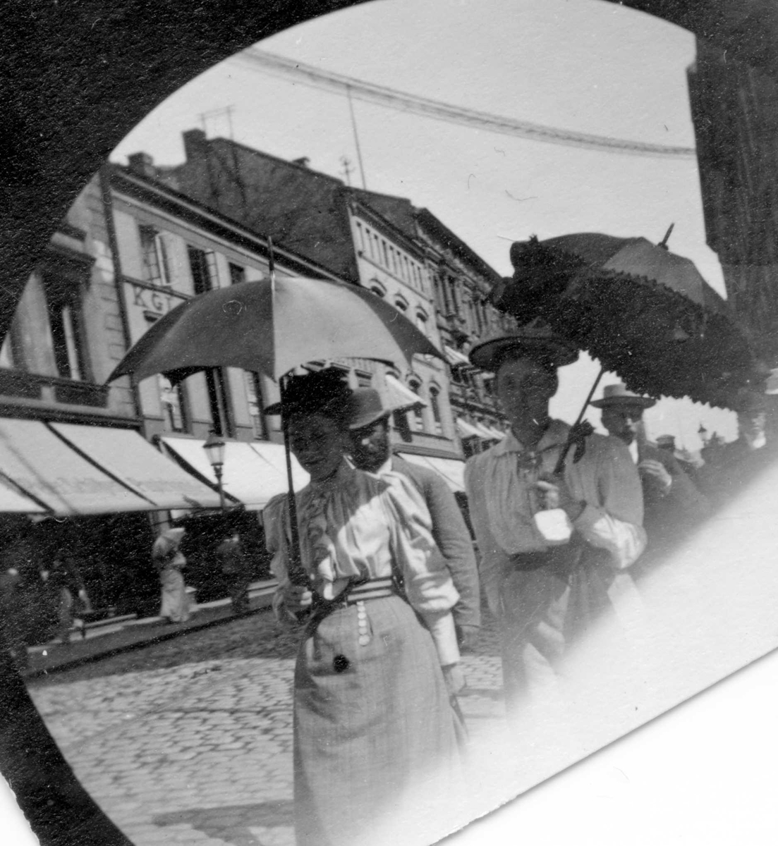 To kvinner med parasoll i bygate, Karl Johans gate, Oslo. Bygårder og butikker bak.