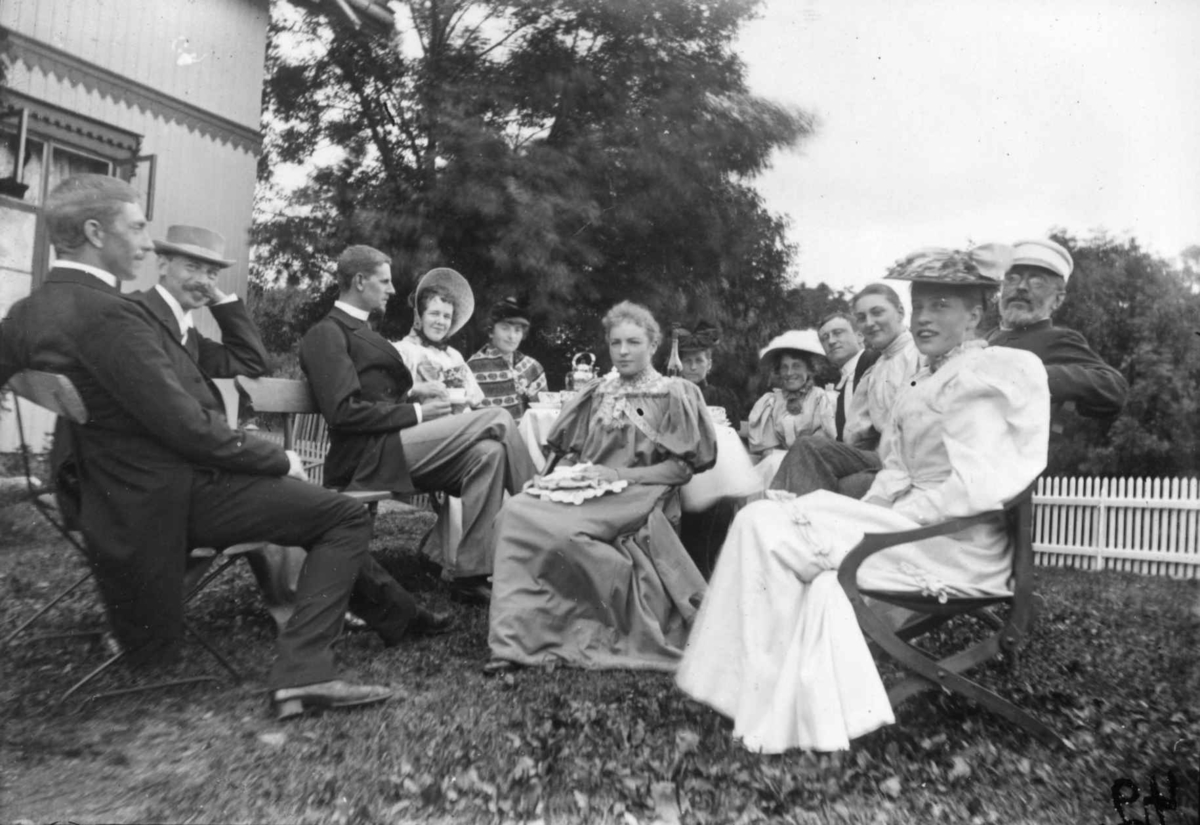 Div. drakt, Norge 1908-10. Kvinner og menn sittende i en  hage.  Mennene i dress. Kvinnene i lange kjoler. Noen med hatter og luer.