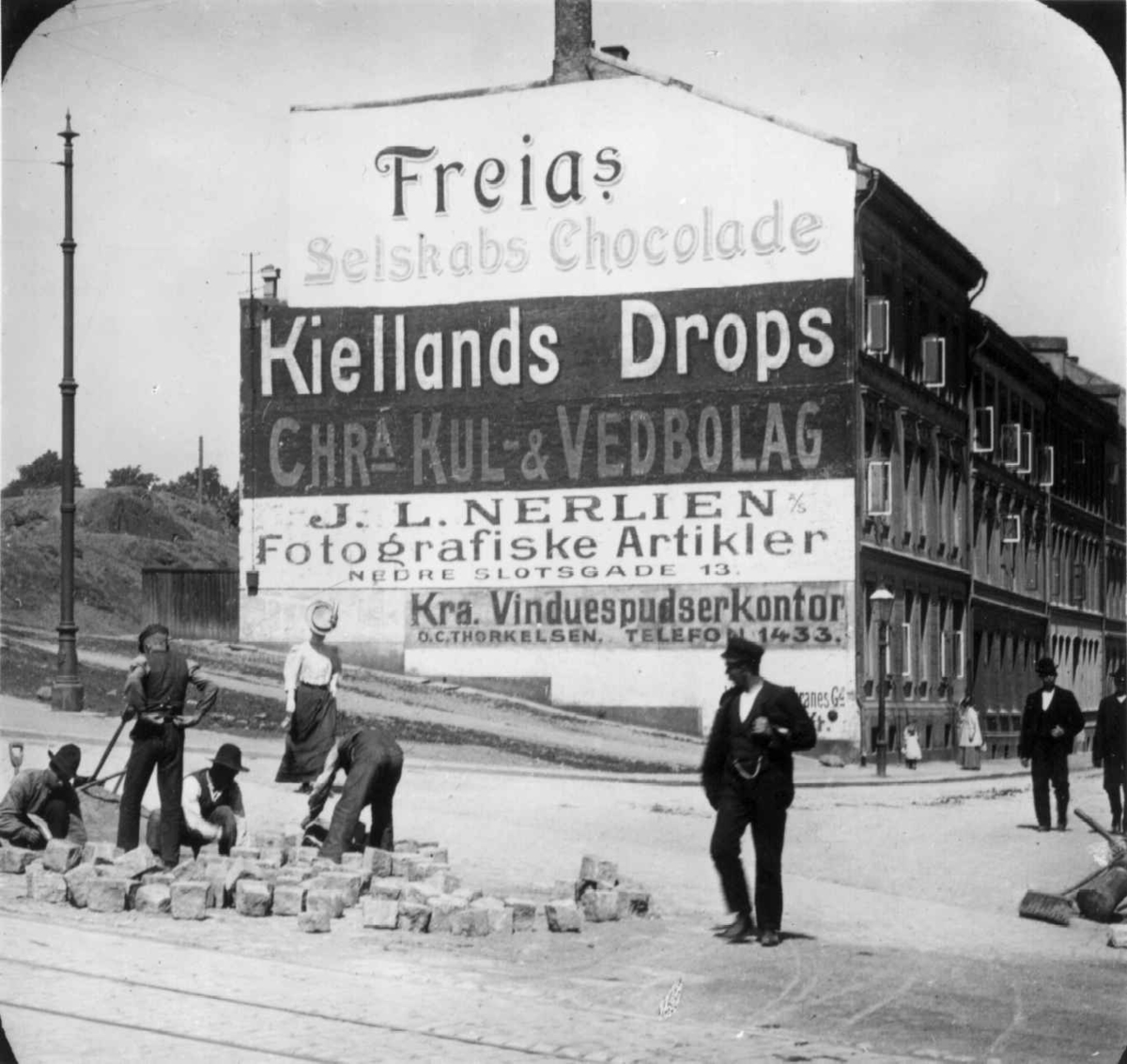 Fra Galgeberg eller ved Ullevålsveien i Oslo. Arbeidere jobber med brostein, vegg med reklame. Til 1905-utst. NF.