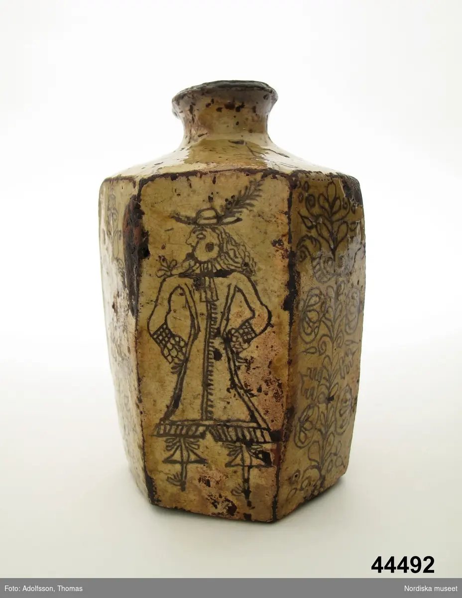 Sjusidig flaska av lergods, engoberad, med ristad dekor med man i 1600-talsdräkt och växtornament.
Se Bonge-Bergengren 1994, s 23.
