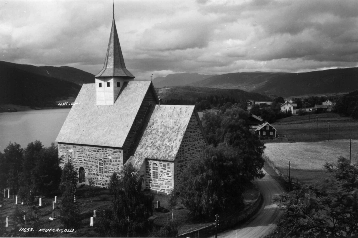 Vestre Slidre kirke, Oppland 1934. Steinkirke bygget 1150-1200. Omgitt av kirkegård med vei forbi. Utsikt mot Slidrefjorden og jordbruksbygd med gårdsanlegg. Åser og fjell i bakgrunnen.