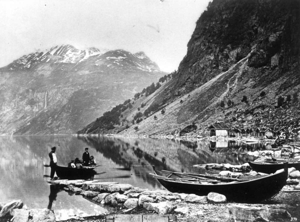 NR. 374 (13,5X18) 
Merok, Geiranger. Et følge av mennesker som er ute i robåt på fjorden.
bebyggelse og båter i strandlinjen, det hele er omkranset av fjell.
