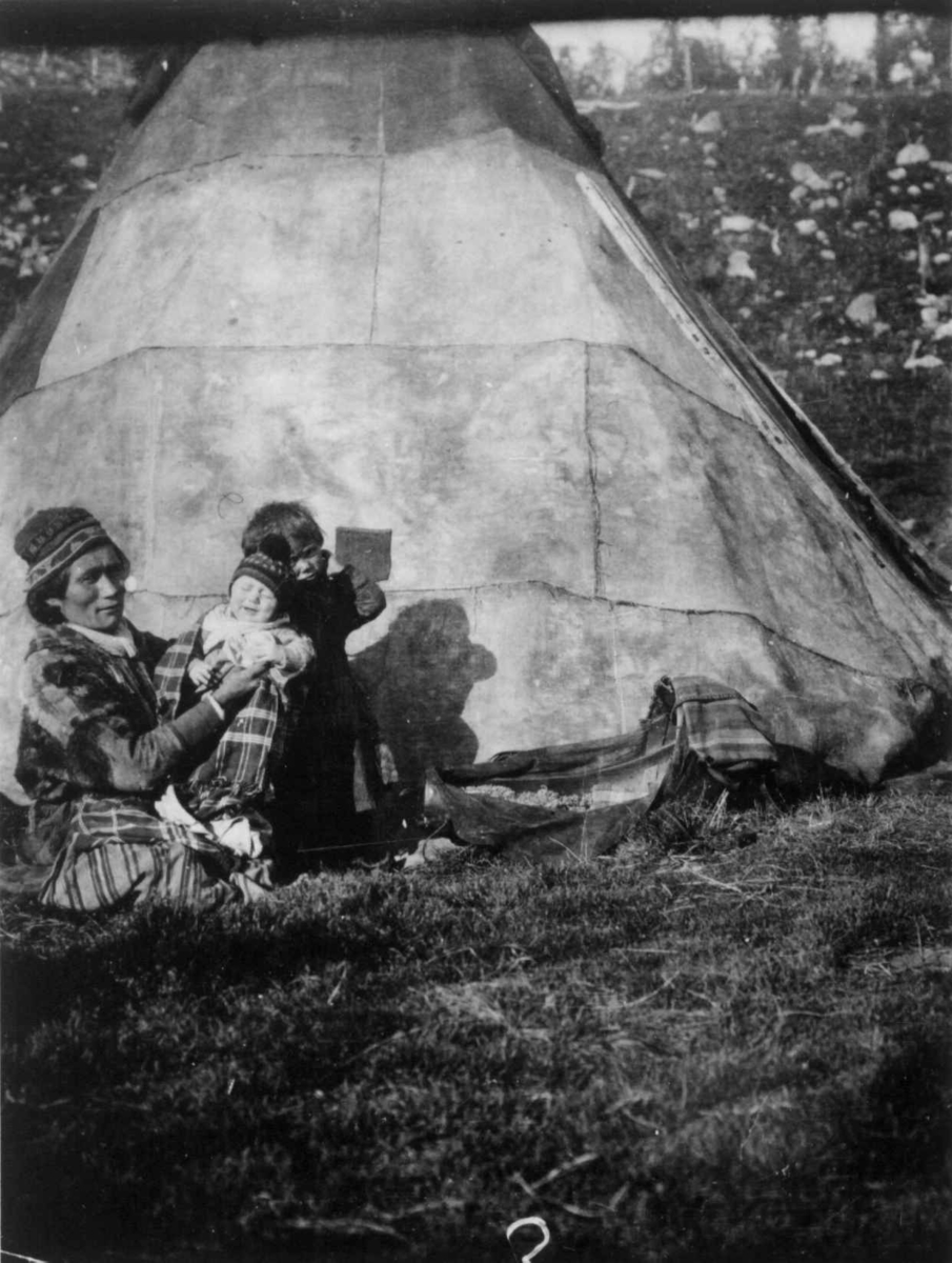 Samekvinne og to barn utenfor teltet, komsen ved siden av er antagelig fylt med mose. Pålnoviken 1914.