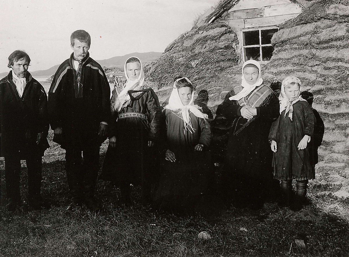 Familiegruppe (6) i samisk drakt, oppstilt foran gamme, Laksefjord, Lebesby. Kvinnen som sitter, nr. 3 fra h., er telegrafistinne, omgitt av sin familie.
Del av serie fra en forskningsreise i Øst-Finnmark 1909.