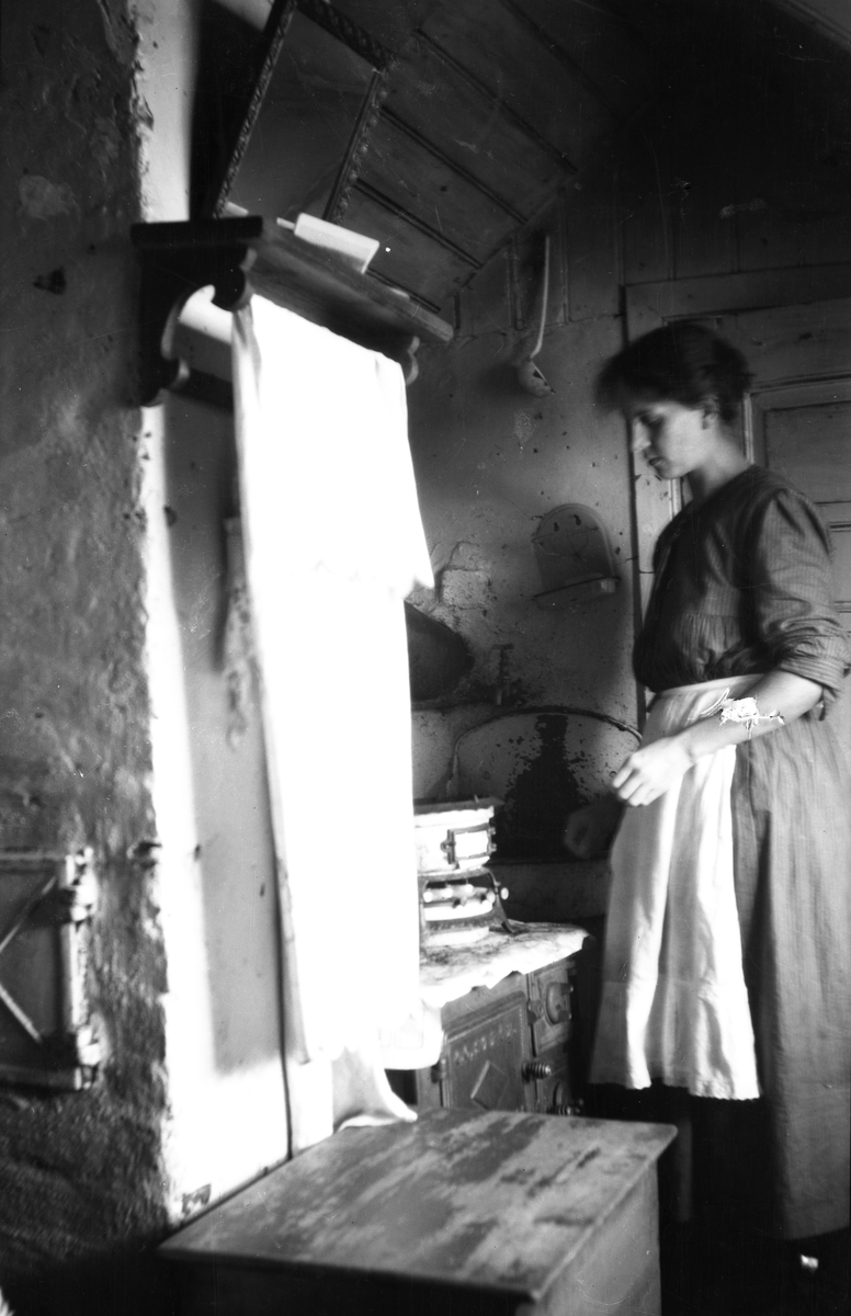 Interiør, kjøkken, Oslo. Kvinne foran komfyr, vedkasse og oppheng med pyntehåndkle i forgrunnen.
Fra boliginspektør Nanna Brochs boligundersøkelser i Oslo 1920-årene.