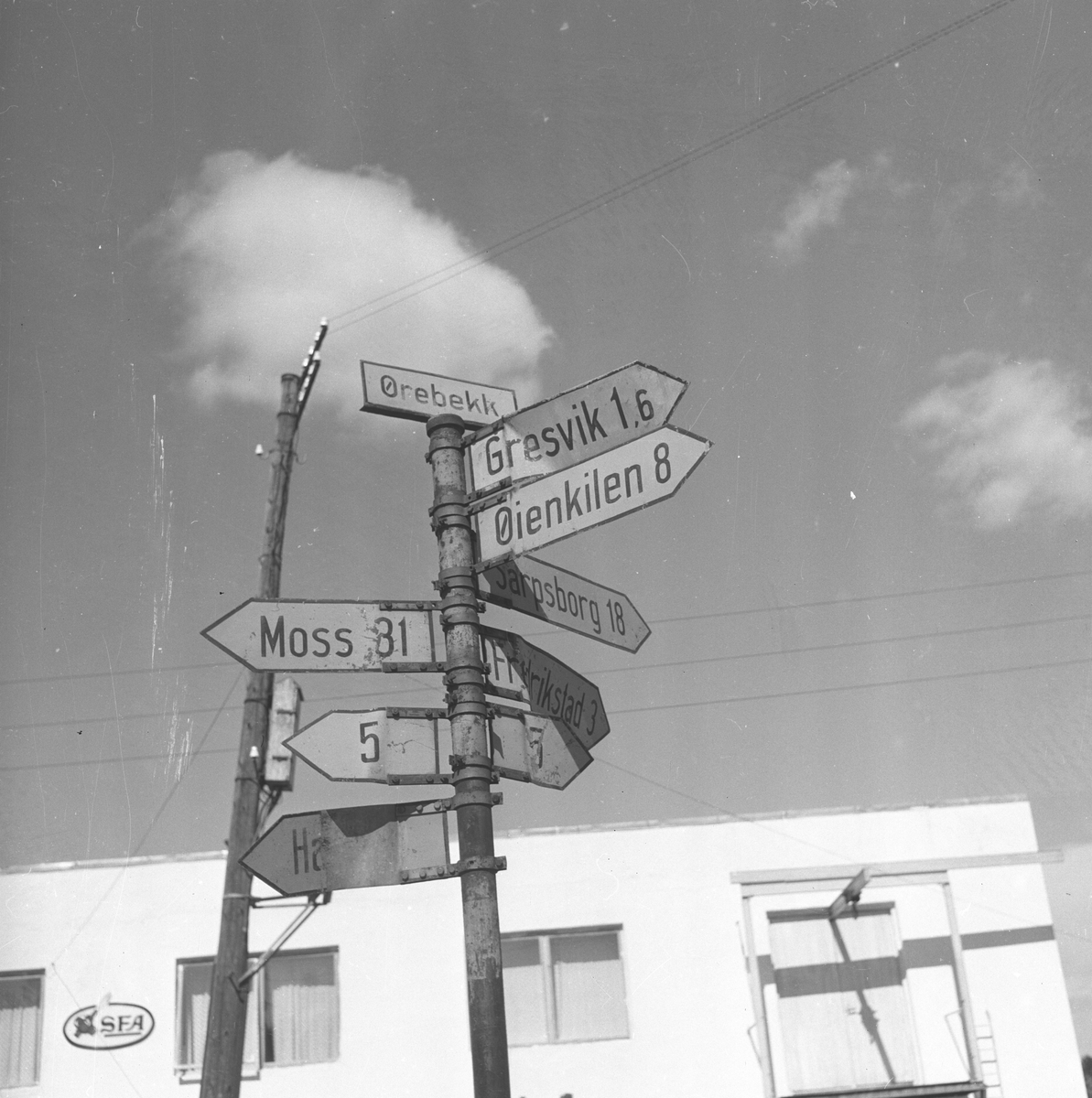 Langs Riksvei 1 ved Ørebekk i Østfold står et skilt med henvisninger til Fredrikstad, Sarpsborg, Moss etc. Fotografert 7. juli 1958.