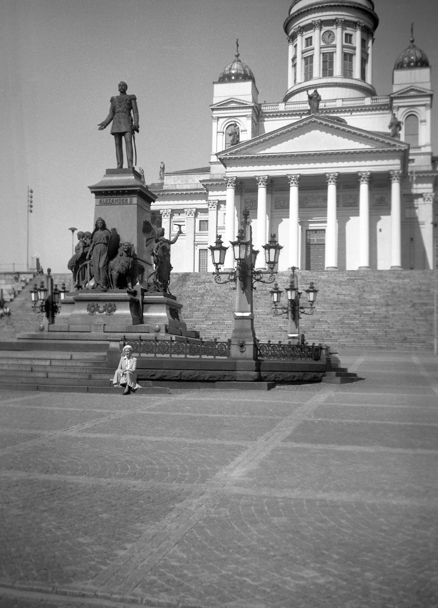 Dordi Arentz sitter ved statue av tsar Alexander 2. foran domkirken i Helsinki. Fotografert 1951.