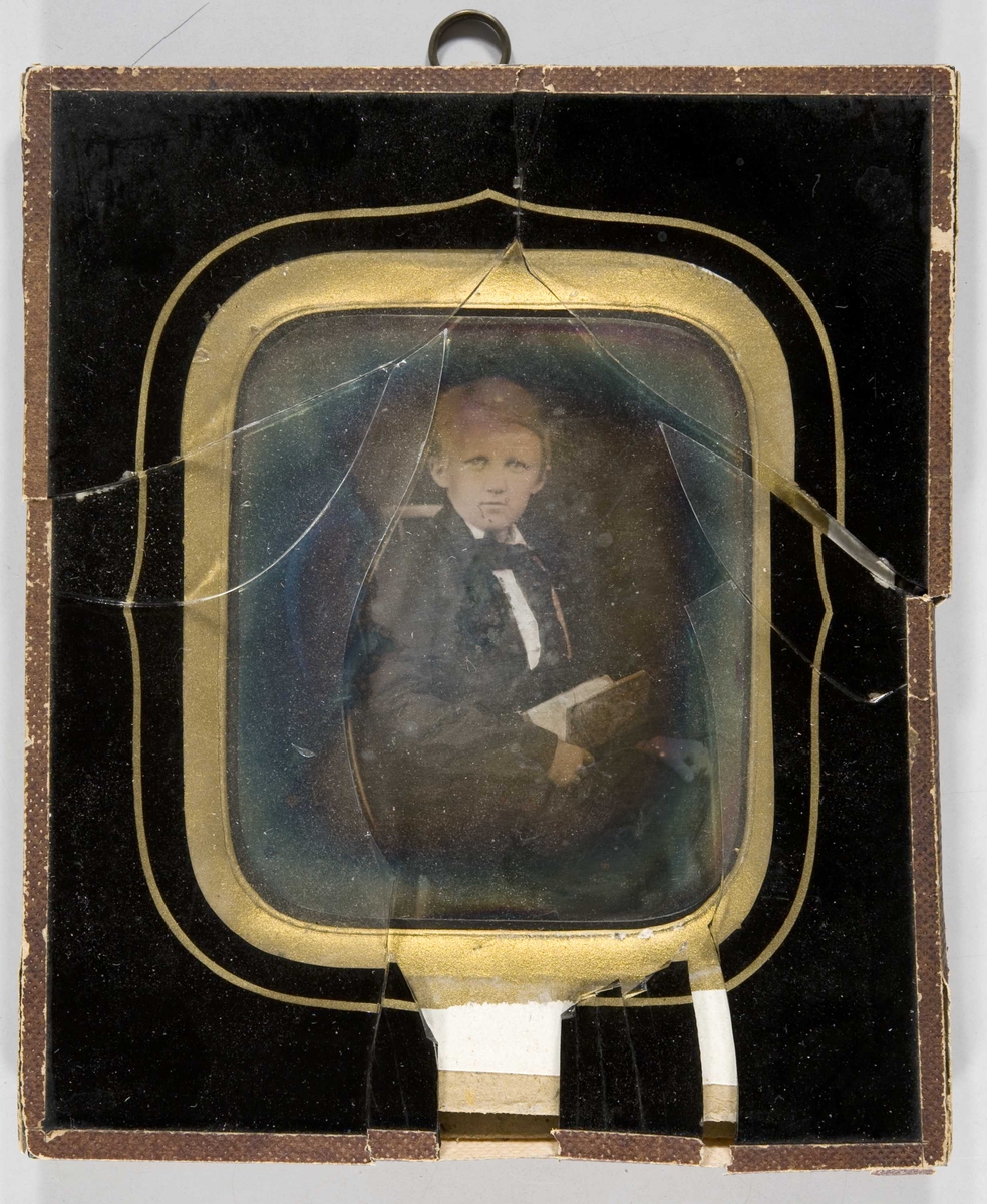 Daguerreotypi. Halvfigur portrett av ukjent gutt. Han sitter med en bok i fanget. Knust glass