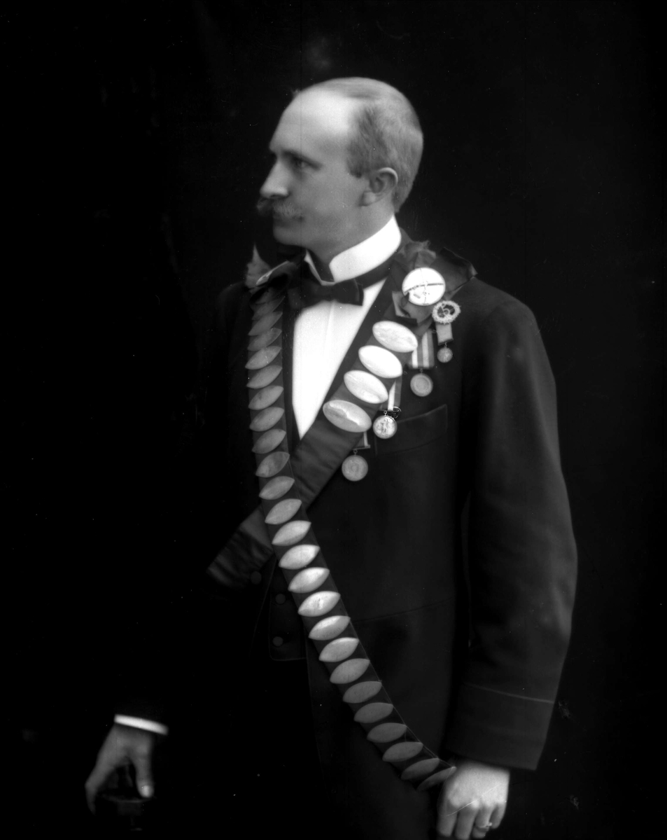Portrett, mann med bånd med premieplaketter, skyting. Kongebeltene til Skydeselskapet Skarpskytten. Ingeniør P Olsen, vinner i 1904(?).