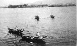 Bilde av båter i vannet med stående roere. By i bakgrunnen. 