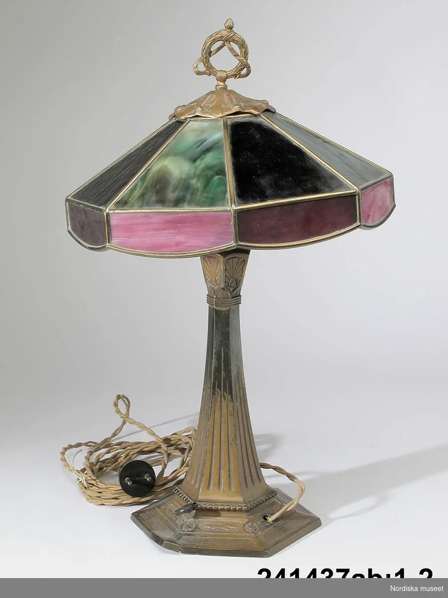 Huvudliggaren:
"Bordslampa, med elektrisk installation; fot av brons; skärm av gröna och röda glas, infattade i gulmetall; spröjsar. Två lampor. Jugendstil."