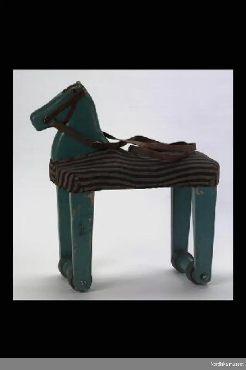 Inventering Sesam 1996-1999:
L 35  B 16 H 39 (cm)
Grönmålad leksakshäst av trä med rullar klädda med läder. Kroppen stoppad och klädd med randigt brunt bomullstyg, tömmar av läder.
Ett litet barn kan sitta på hästen och sparka sig fram. 
Birgitta Martinius 1996