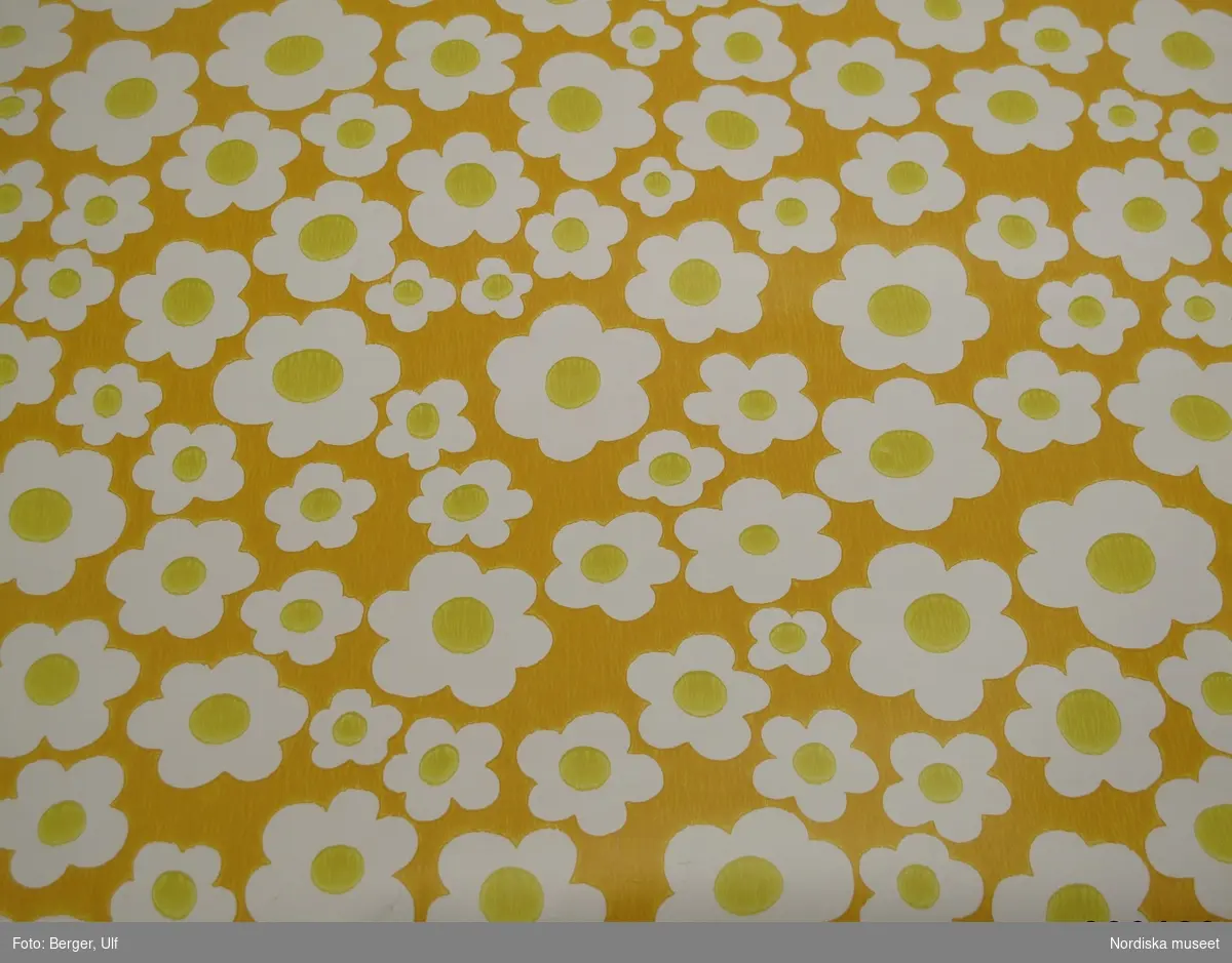 Tapetprov, del av tapetrulle. papper, tryckt mönster, enkla vita blommor med gul mitt mot gul botten. Inga stämplar på baksidan. Kantskuren.
/Leif Wallin 2010-07-09