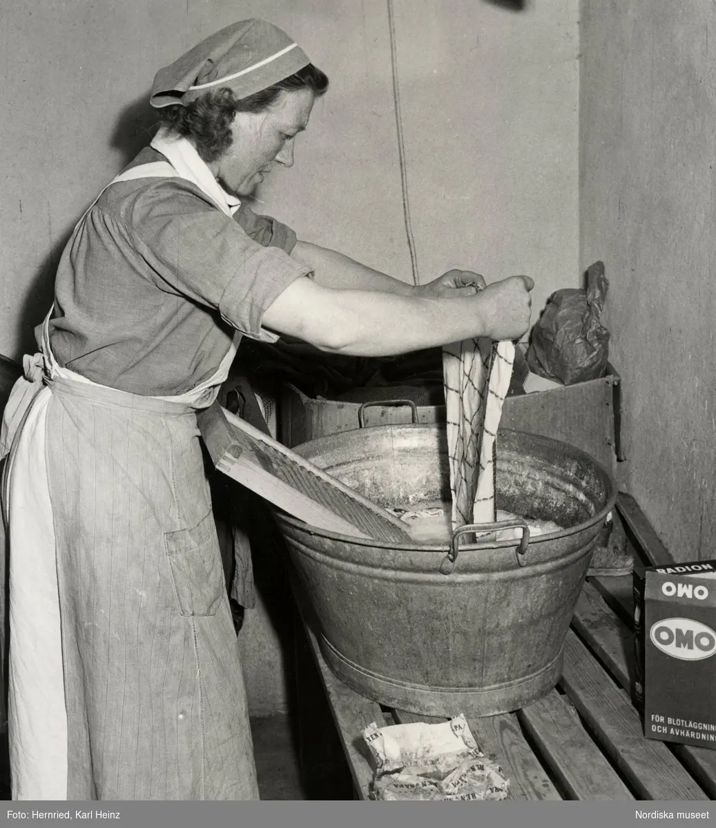Kvinna tvättar i zinkbalja med tvättbräda. Tvättmedel är av fabrikaten Ren Såpa, Radion och OMO.
