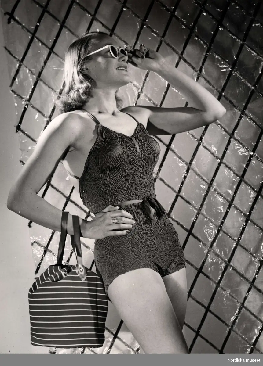 Dammode från varuhuset Nordiska Kompaniet i Stockholm 1944. En modell poserar i krinklad baddräkt med knytskärp i midjan.Hon bär solglasögon och över armen hänger en liten strandväska.