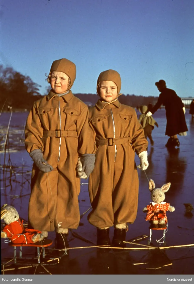 Barn med leksaker ute på is.Två flickor i overaller står på isen med varsin leksakskanin på små kälkar.