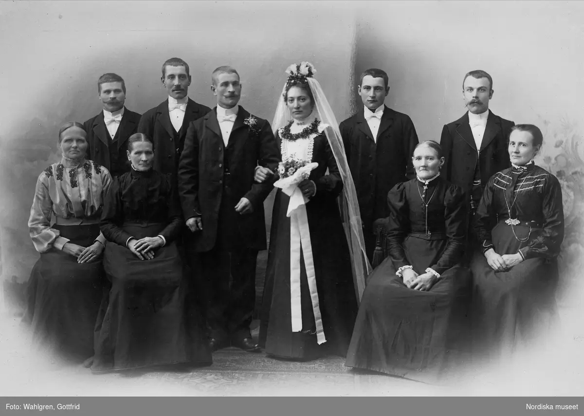 Bröllop, Tranås. Gruppbild med brudpar stående i mitten. Bruden iklädd svart klänning och vit slöja. Brudbukett och blomsterkrans runt halsen.