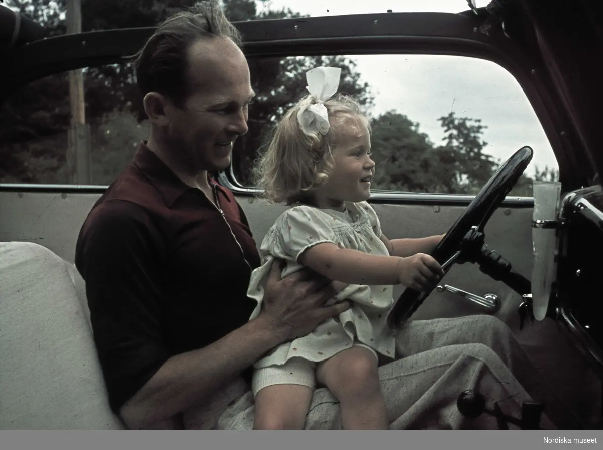En man sitter i en bil med en liten flicka i knät som håller i ratten.