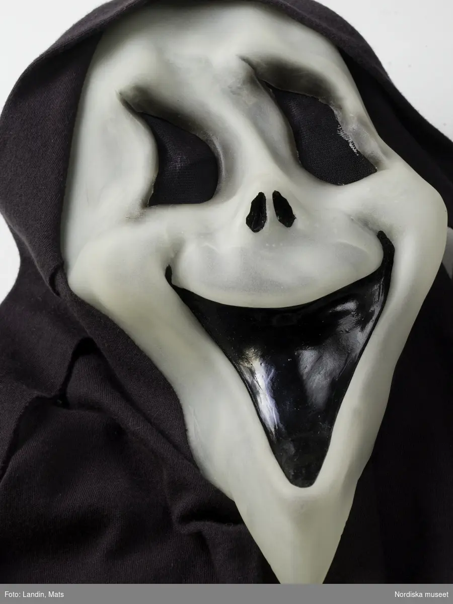 Liemansmask, halloweenutklädsel för barn, ansiktsmask av halvgenomskinlig vit plast med svart mun, ögonhål med svart nät, infälld i lång huva av svart bomullstrikå, hängande flikar vid öronen. Nordiska museets föremål inv.nr 328022.