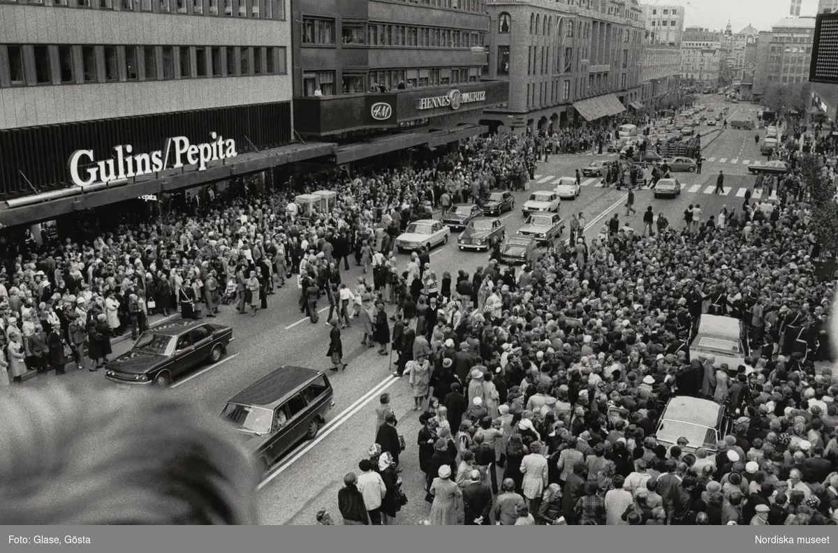Hamngatan utanför Gallerian köpcentrum, Stockholms första gågata under tak. Ett myller av människor närvarar vid invigningen 7 oktober 1976. Poliser dirigerar trafiken och håller folksamlingen tillbaka.