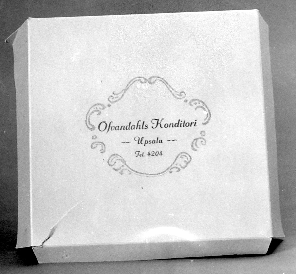 Tårtkartong av vit papp med uppfällbart lick,  blått tryck  "Ofvandahls konditori,  Upsala tel 4204".  Locket gar två breda flikar och en smal.
