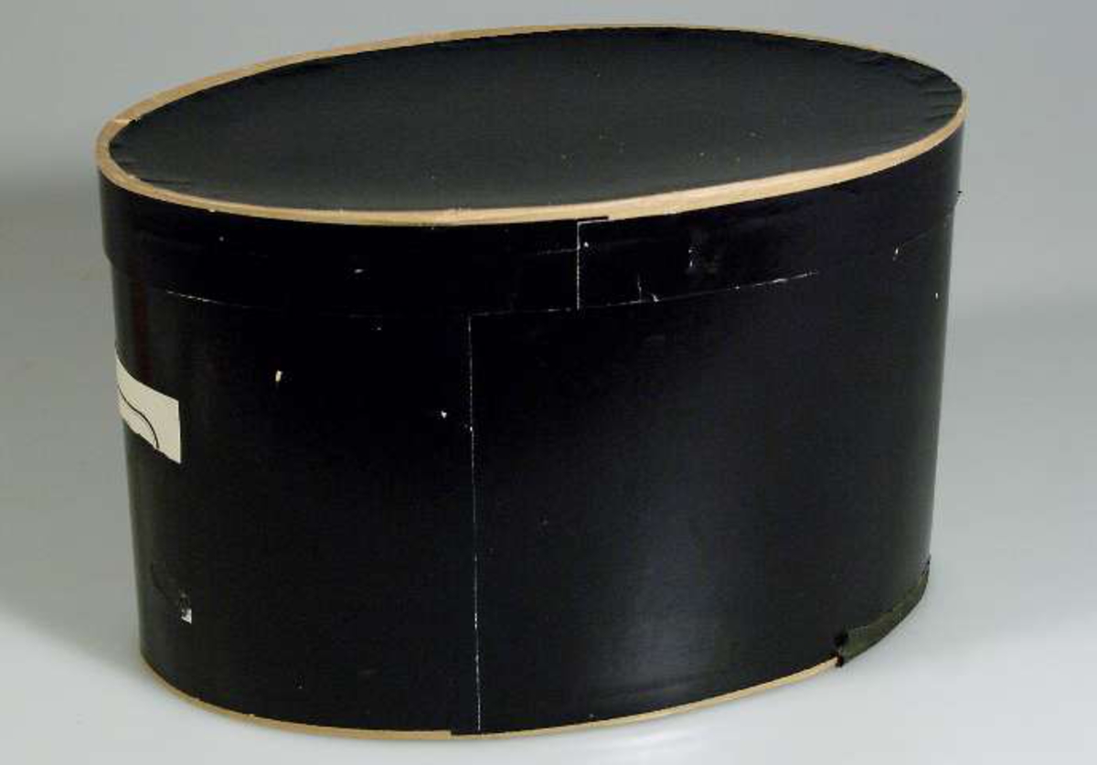 Oval ask med lock, av papp, klädd med svart papper.
Innehåller krimmermössa UM29562. 
