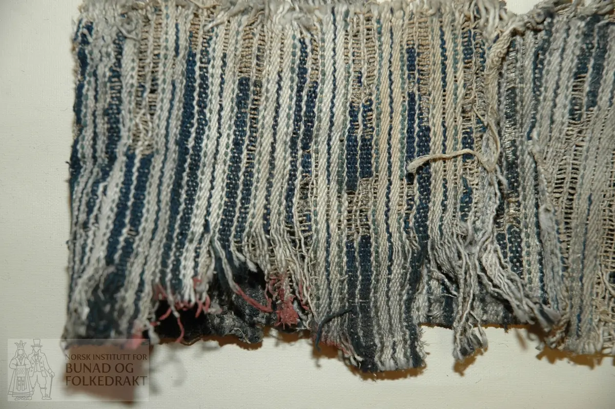 Nedre del av eit buksebein. Renning av ubleika lin, innslag av mørk blå og mellom blå ull, samt ubleika bomull.