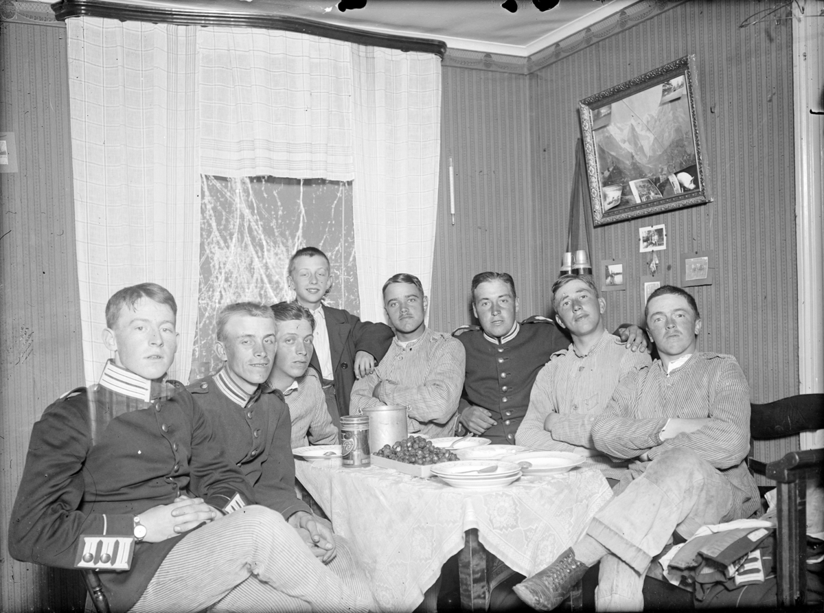 Grupporträtt. Sju män och en pojke samlade vid ett matbord. Tre av männen är iförda arméuniform från I 4 eller I 5 regemente. På bordet står tallrikar och ett fat köttbullar. Mannen längst till vänster är Ernst Axfors.