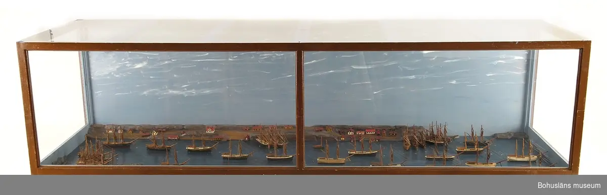 Panorama i monter över Fiskebäckskils hamn på 1880-talet med 33 fartygsmodeller i vinterkvarter.
Tillverkad av kapten John Emil Olsson (1880-1950), Fiskebäckskil på Skaftö i Lysekils kommun 1942.
Trä, metall, lintråd, gips, glas, målat med oljefärg.
Största fartygsmodell längd 16 cm; monter 202 x 61 cm, höjd 53 cm; okänd skala. Vattenlinjemodeller av 30 råseglare, två större jakter och en galeas, uppankrade eller förtöjda. Stående och löpande rigg, däckshus och lastluckor, målade och lackade placerade på en blåmålad platta. Bakgrund av utskurna och målade berg med ett 30-tal hus, sjöbodar och bryggor utspridda längs den djupgående kilen och med gatten ut mot Gullmaren.

Framtill på modellens långsida står textat med vita tryckbokstäver: 
FISKEBÄCKSKIL:S HAMN I SLUTET AV 1880-TALET.
VID VARJE FARTYGS FÖR STÅR EN SIFFRA SOM HÄNVISAR TILL TAVLAN OVANFÖR MONTERN.
Den maskinskrivna texten berättar:
Fiskebäckskils hamn i slutet av 1880-talet. 
I vinterkvarter upplagda i Fiskebäckskil hemmahörande segelfartyg.
1. Skonert Gustaf, befälhavare M. Johansson
2. Brigg Clinton, befälhavare J. Bengtsson
3. Skonertskepp Nordstjernan, befälhavare Am. Olsson
4. Barkskepp Diana, befälhavare H. Andersson, Gåsö
5. Skonert Elise, befälhavare Joh. Gren
6. Barkskepp Celeritas, befälhavare C. Bengtsson
7. Brigg Ben:s Mollén, befälhavare Joh. Månsson
8. Skonerter Elida, Magnus Gren
9. Skonert Olga, befälhavare C. Jansson
10. Skonert Idun, befälhavare Ferd. Gren
11. Skonert Ida Sofia, befälhavare O. Simonsson
12. Skonert Gustaf, befälhavare J. Andersson
13. Skonert Alfa, befälhavare A. Kihlman
14. Skonert Klara, befälhavare P. Berntsson
15. Jakt Tärnan, befälhavare P. Persson
16. Barkskepp Bohuslän, befälhavare H. Jonasson
17. Galeas Blenda, befälhavare B. Wahlberg
18. Jakt Wiktor, befälhavare J. Hansson
19. Brigg Gerda, befälhavare F. Edman, Flatön
20. Skonert Norma, befälhavare A. Larsson
21. Brigg Gustava, befälhavare J. Larsson
22. Skonertskepp Uddevalla, befälhavare J. Mattsson
23. Skonert Carolina Matilda, befälhavare C. Kihlman 
24. Skonert Charlotte, befälhavare Osk. Olsson
25. Briggen Swartvik, befälhavare J. Berntsson
26. Skonerten Sirius, befälhavare A. Ödman, Ögården
27. Briggen Agnes, befälhavare P. Larsson 
28. Skonert Sofia, befälhavare A. Karlson, Gåsö
29. Skonertskepp Carl, befälhavare Sim. Andersson 
30. Skonert Anni, befälhavare H. Nilsson 
31. Skonert Amfitrite, Kilhlberg
32. Brigg Mary, befälhavare C. L. Kristensson 
33. Skonert Alfhild, befälhavare Osk. Bengtsson
Anm. Nr 1? (svårtydd siffra) och 25 förliste i Nordsjön maj månad 1895.
Nr 21 kolliderade i Engelska kanalen 1890. Blev räddad och reparerad.
Nr 5 förliste i Nordsjön hösten 1903 med man och allt.
Nr 19 ligger som museum i Gevle hamn. De övriga fartygen har tidens tand gnagt sönder, antingen här eller på främmande ort.

Definitioner: 
Råseglare
Segelfartyg som huvudsakligen är riggad för råsegel t.ex. fullriggare, bark el. brigg.
Råsegel - rektangulärt eller trapetsformigt segel som förs på en rå, dvs. en horisontell stång fäst vid mastens framkant. 
Bark
Segelfartyg med tre eller fler master med rår på alla master och ett gaffelsegel på den aktra.
Råsegel - se ovan. 
Gaffelsegel - trapetsformat segel fäst längs akterkanten av masten och med sina övre (kortare) och undre (längre) lik (kanter) fäst vid två rundhult (gaffel resp. bom).
Brigg 
Tvåmastat segelfartyg med rår på bägge masterna och ett gaffelsegel på den aktra.
Råsegel - se ovan. 
Gaffelsegel - se ovan.
Skonertskepp
Segelfartyg med tre eller fler master av vilka den förligaste har fullständig råsegeltackling och gaffelsegelrigg på de övriga. Kallas också barkantin.
Skonert
Segelfartyg med två eller fler master av vilka den förligaste har fullständig råsegeltackling och övriga gaffelsegel med trekant-toppsegel.
Råsegel - se ovan. 
Gaffelsegel - se ovan. 
Galeas
Ett mindre, tvåmastat segelfartyg riggat med gaffelsegel som skonare med stormasten främst. 
Gaffelsegel - se ovan.
Jakt
Enmastat segelfartyg med öppet däck och en mindre kajuta akterut. 
Gaffelsegel - se ovan.