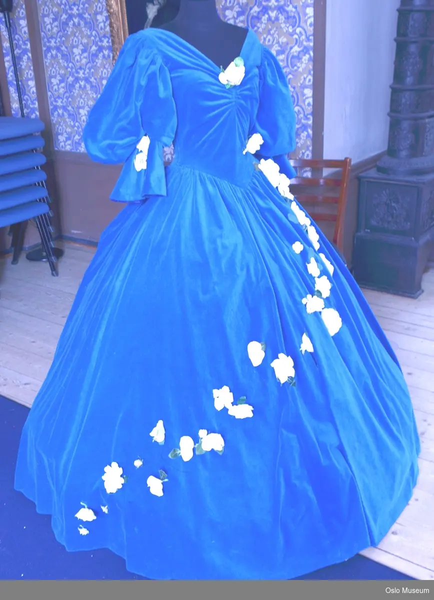 Lang kjole i blått fløyelsstoff med halvlange pose-ermer, dekorert med påsydde hvite blomster i stoff. Tilhørende underskjørt.