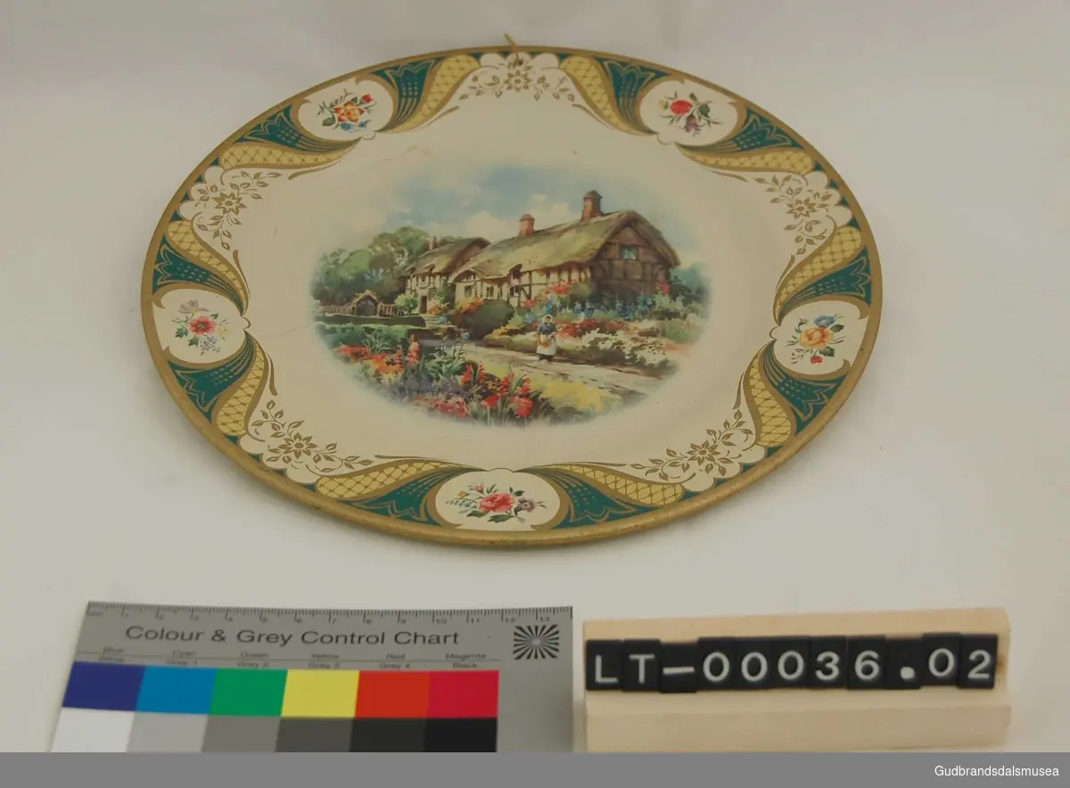 To tallerkener i blikk. Dekorert med border og blomsterornamentikk rundt kanten, maleri av hage med to bindingsverkhus og en kvinne i midten.