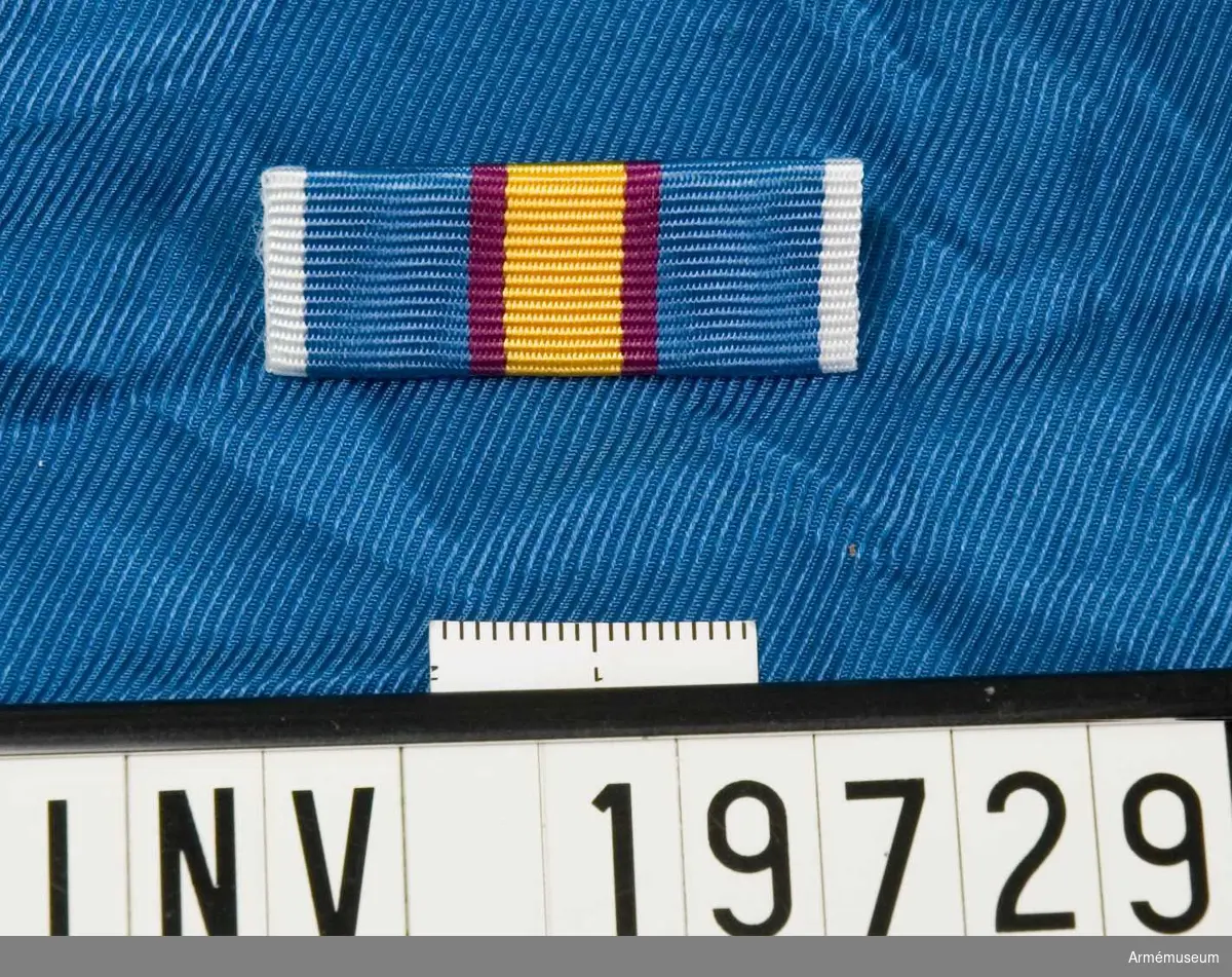 Blått band med vita kanter och en bred gul rand i mitten åtföljd av en purpurröd rand på vardera sidan. Släpspännet förvaras i ask tillsammans med en medalj och miniatyrmedalj.