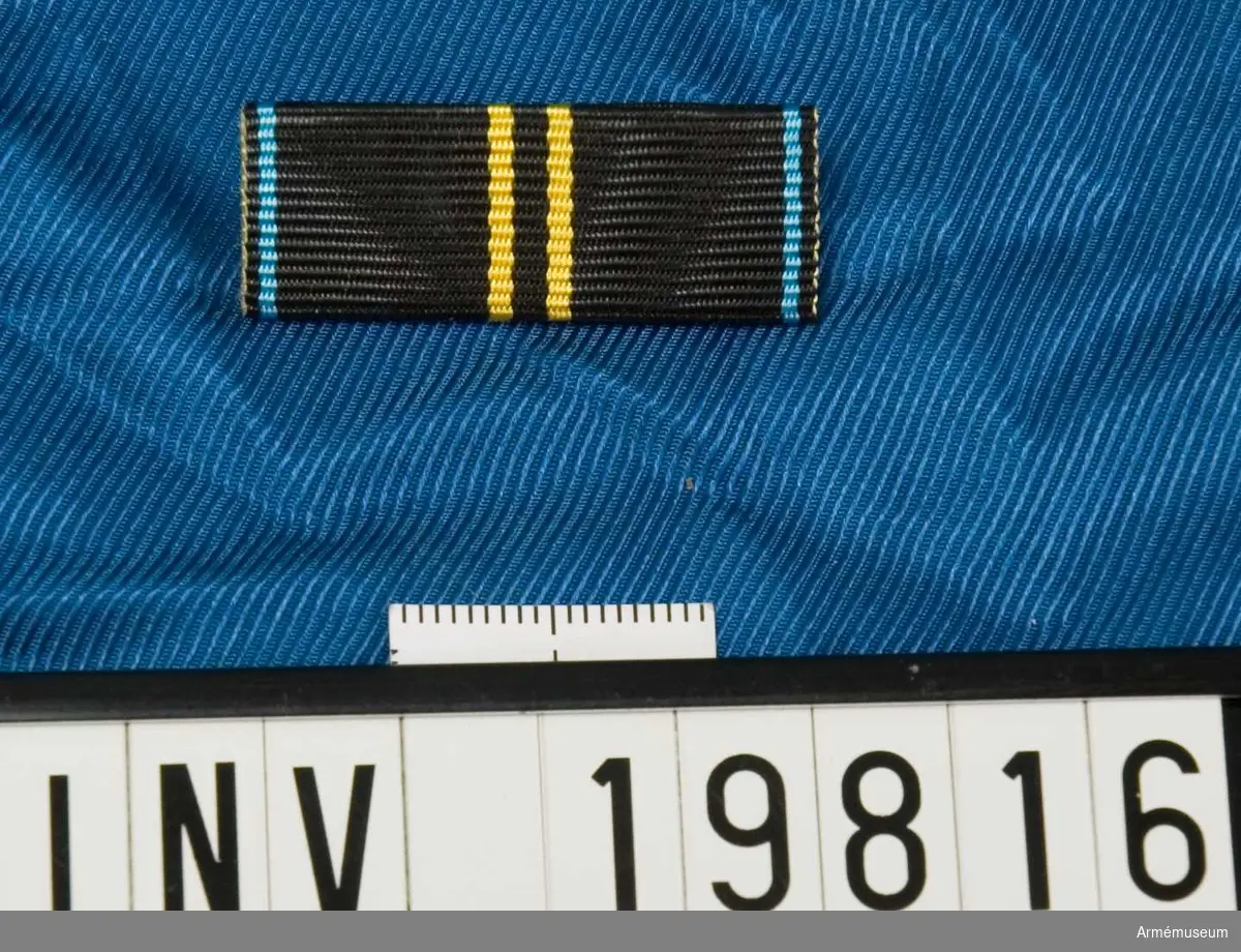 Svart band med två gula streck på mitten och ett blått streck på vardera kanten. Släpspännet förvaras i en ask tillsammans med en medalj och en miniatyrmedalj.