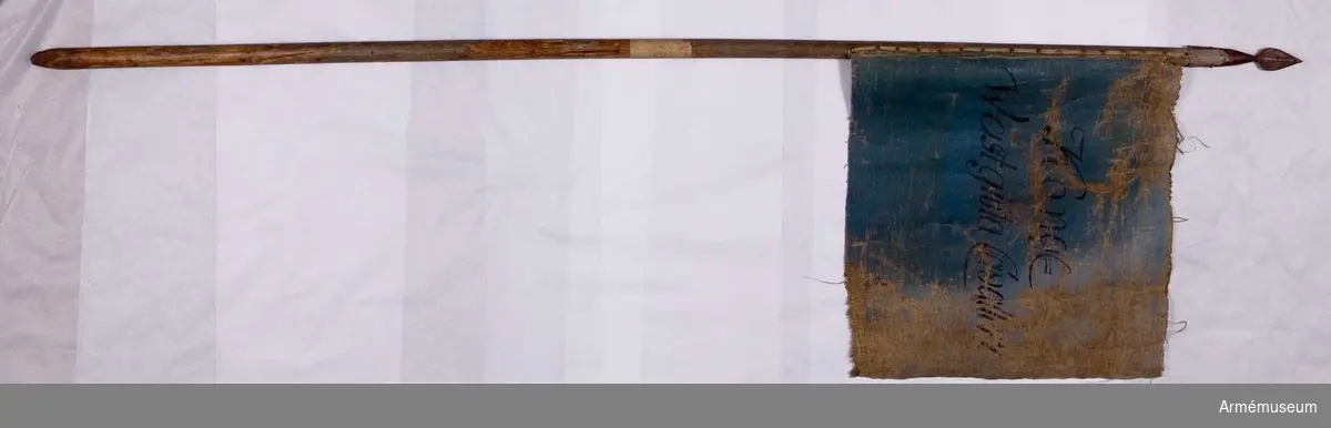 Duk: Tillverkad av enkel blå och gul lärft. Dukens vertikala ytterkant stad. Fäst vid stången med mässingsspikar genom dubbelvikt duk utan att lindas runt stången.

Dekor: På inre sidan målad blå med inskrift i svart på två rader.

På yttre sidan målad gul med inskrift i svart på två rader.

Stång: Tillverkad av gråblåmålad furu. Spets med holk av järn. Spetsen har lansettformat blad.
