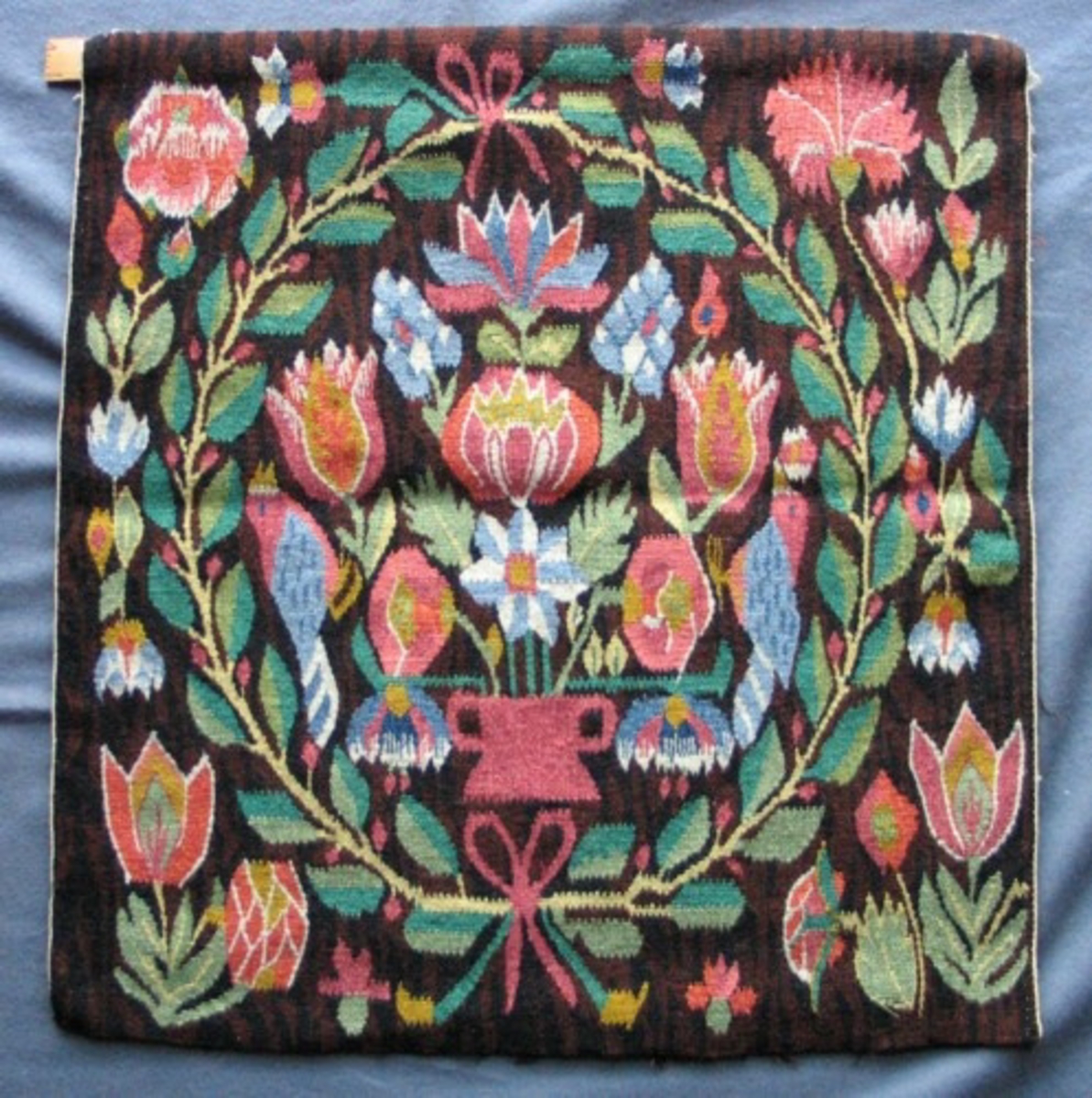 Flamskvävnad med motiv av en röd urna med en stor blomsterbukett flankerad av två blå fåglar i bladkrans. Olika blommor utanför kransen, större i hörnen. Något dämpad färgskala mot brunsvart botten. Varpen knuten med langett och därefter flätad mot baksidan där trådarna är nersydda två och två. Monterad att hänga.
