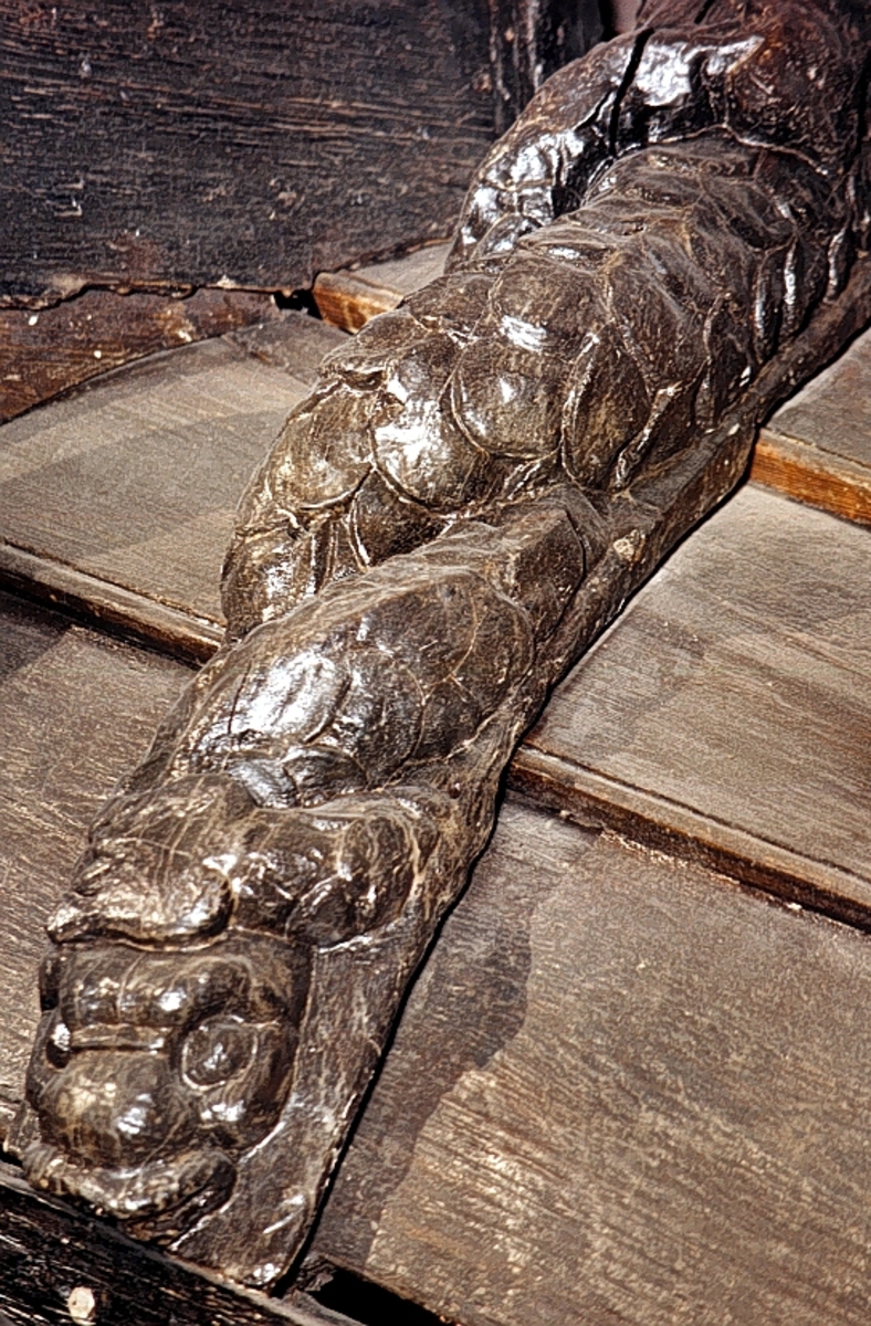 Triton med mustascher och tjockt, vågigt hår. Armarna är reducerade till upprullade bladflikar. Runt midjan sitter ett band med en oval medaljong i mitten. Skulpturens fiskstjärt slingrar sig kring stjärten till en fisk eller delfin, vars huvud är synligt vid skulpturens fotända.



Bakstycket är kraftigt och snidat i ett med skulpturen. Baksidan är försedd med hak eller urtag. Skulpturens profil är svagt S-formad.

Skulpturen är kraftigt sliten och anletsdragen i det närmaste utplånade.



Text in English: Triton with moustache and thick wavy hair. The arms are replaced by rolled-up leafy decoration. Round the waist there is a girdle or belt with oval decoration. The fish tail is entwined with the tail of a fish or dolphin whose head can be seen at the foot of the sculpture.



The backboard is robust and carved in one piece with the figure. The back side of the backboard was carved with hollowed out grooves. The profile is slightly S-shaped.

The sculpture is badly worn, the features have been practically obliterated.