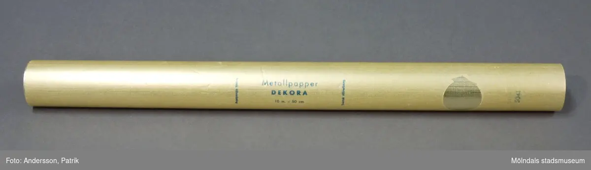 Rulle, "Metallpapper Dekora". Pappret är guldfärgat, ihoprullad i vaxpapper. Storlek: 10 m x 50 cm. Pappret är tillverkat på Papyrus i Mölndal.Litteratur: Papyrus 1895-1945, Minnesskrifter, Esseltes Göteborgsindustrier AB, Göteborg 1945.